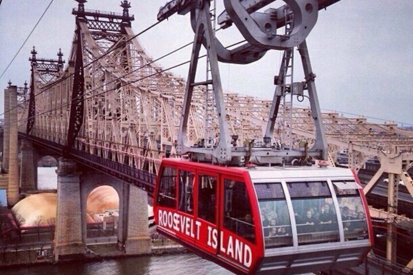 A cable car with 'ROOSEVELT ISLAND' written on it, crossing a bridge with a cityscape in the background. 1