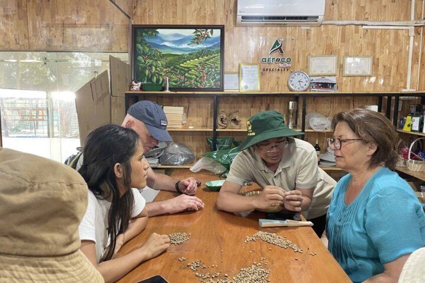 A group of people sitting around a wooden table with scattered seeds, a television displaying a landscape, and shelves with various items and certificates in the background. 2