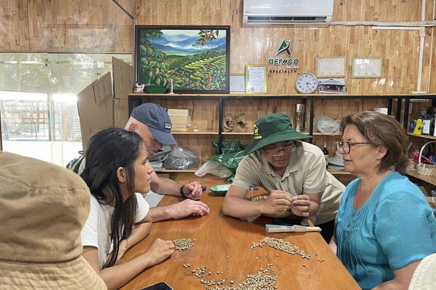 A group of people seated around a wooden table with scattered seeds, a television displaying a landscape, and a shelf with certificates and a clock. 2