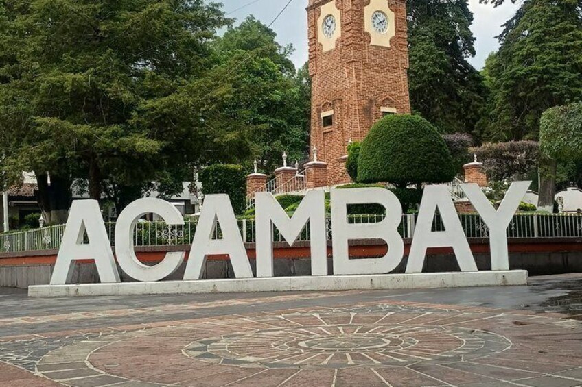 A large sign with the word 'ACAMBAY' in white letters, a clock tower, and a circular pavement design. 1