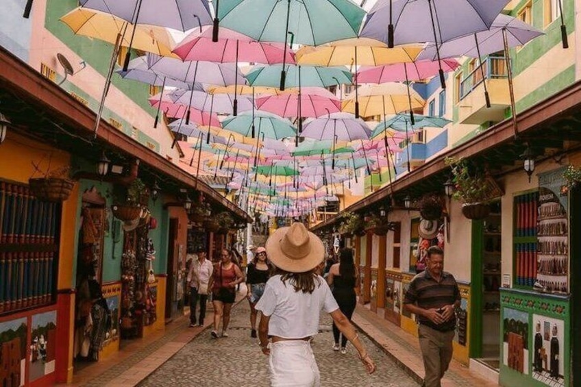 A colorful street with hanging umbrellas, people walking, and shops on either side. 1