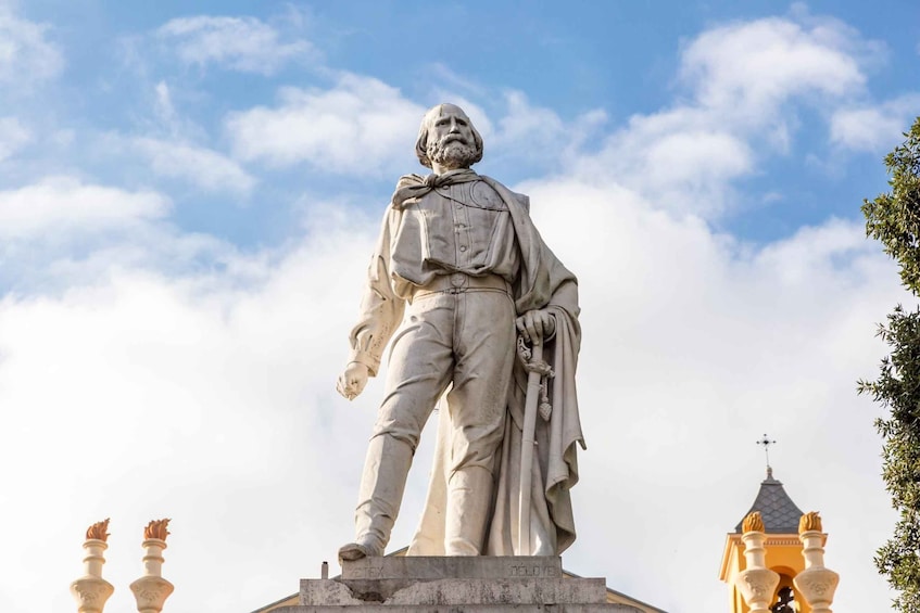A statue of a man in historical attire, standing on a pedestal with inscriptions, set against a backdrop of a clear sky and a building with a distinctive roof. 1