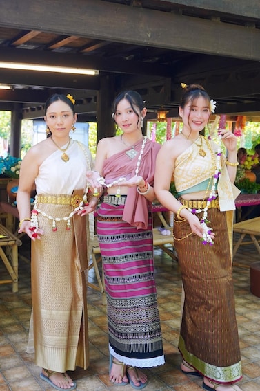 Three women in traditional attire with elaborate necklaces and headpieces, standing under a wooden structure. 1