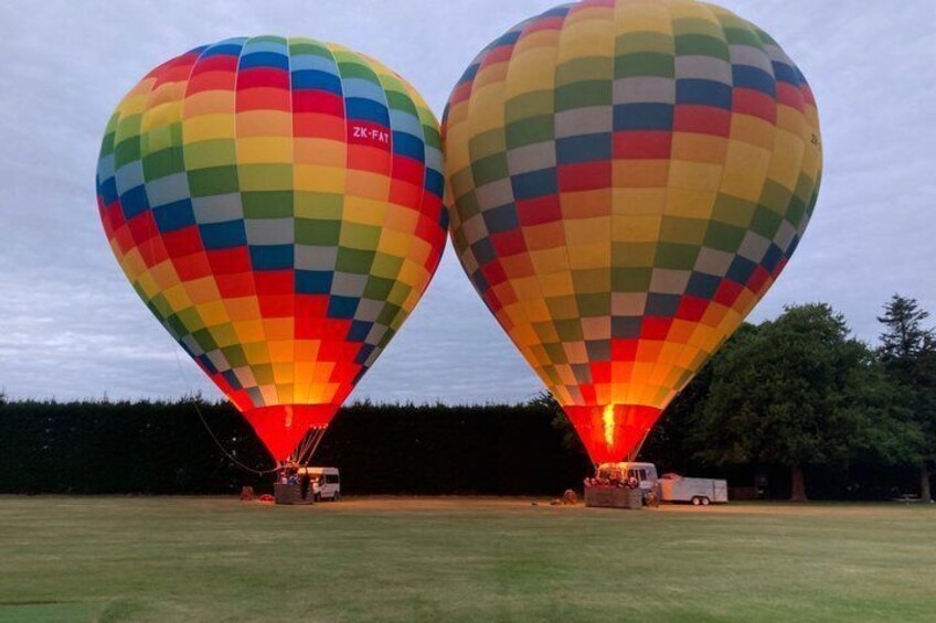 Two hot air balloons with colorful patterns, parked on a grassy field with trees in the background. 2