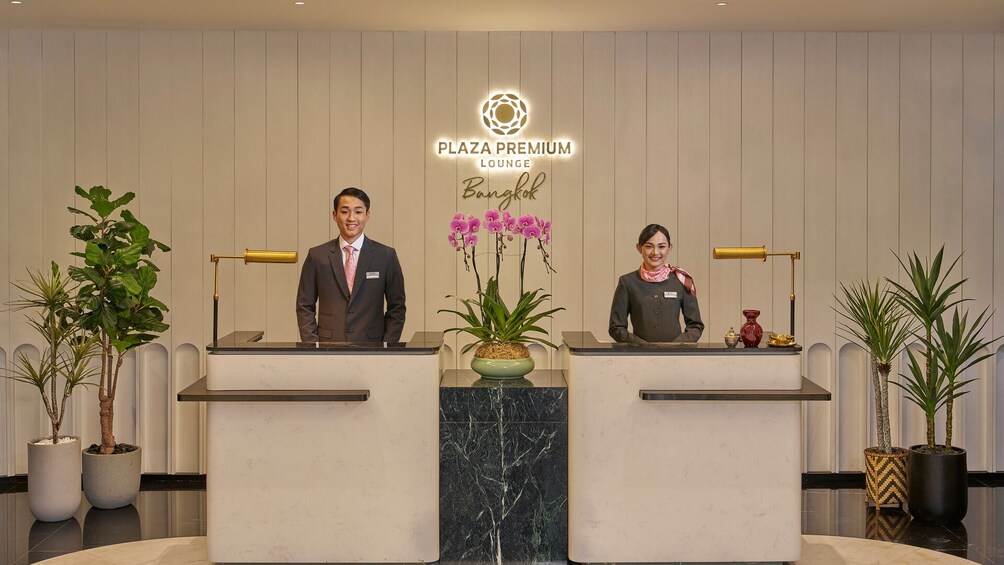 A reception desk at Plaza Premium Lounge in Bangkok with two staff members, potted plants, and a decorative vase. 1