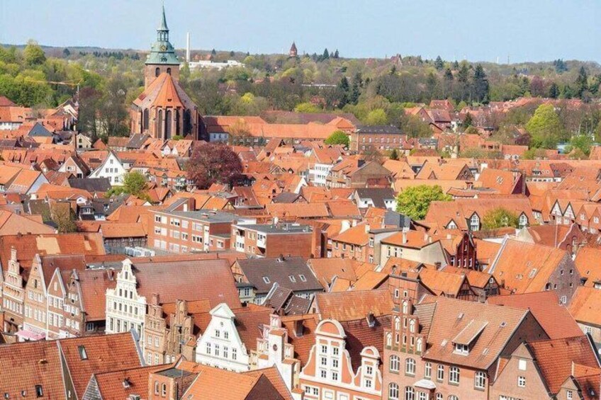 A densely packed town with red-roofed buildings, a prominent church with a spire, and a hilly landscape in the background. 1