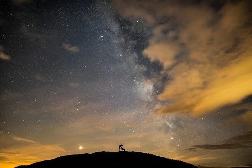 A night sky with stars and the Milky Way, a silhouette of a person on a hill, and a faint moon. 1