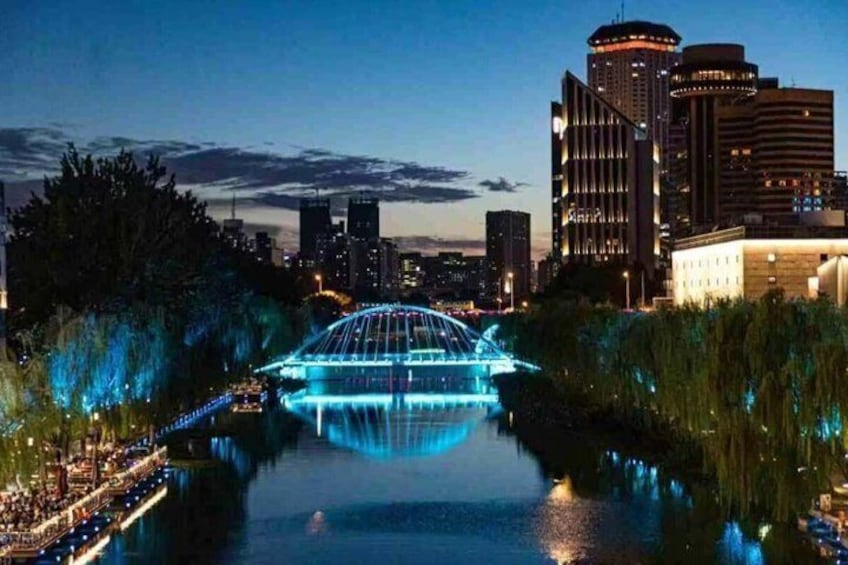 A cityscape with a lit bridge over a river, surrounded by buildings and trees. 1
