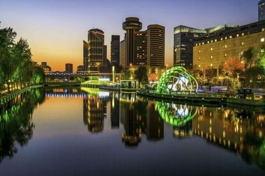 A cityscape at dusk with illuminated buildings, a reflective water body, and a green-lit sculpture. 2