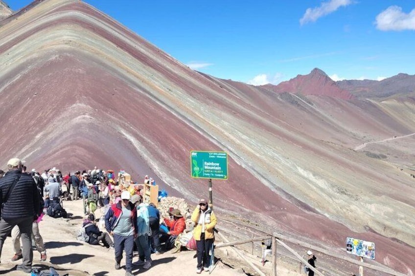 A mountainous landscape with a crowd of people at the base, a signpost, and a clear sky. 1