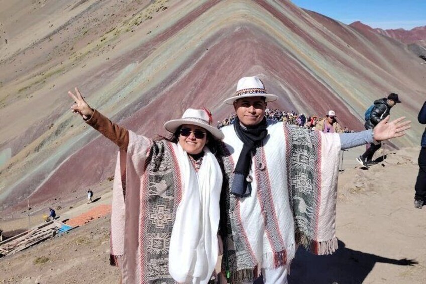 Two people in traditional attire standing in front of a colorful mountain range. 2