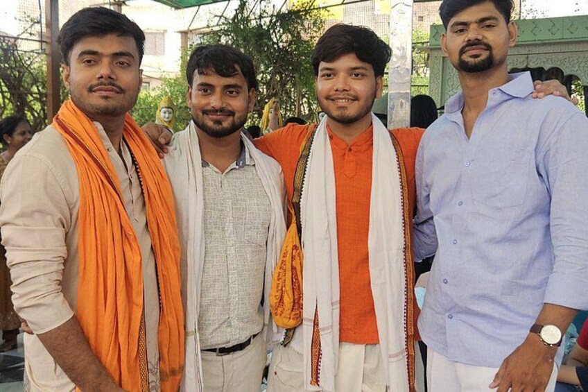 Four men standing outdoors, dressed in traditional attire, with one in a light blue shirt and others in orange and white garments. 3