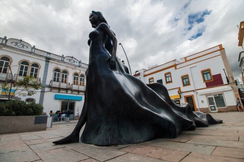A statue of a woman in a flowing dress, a building with arched windows, and a cloudy sky. 1