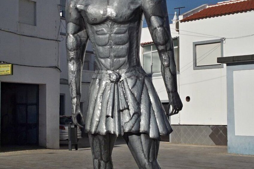 A large metallic statue of a muscular figure wearing a skirt, standing in an urban setting with buildings and a clear sky in the background. 4
