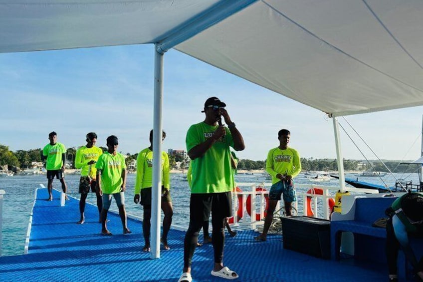 A group of people on a boat with a blue deck, some wearing bright green shirts, and a person taking a photo. 4