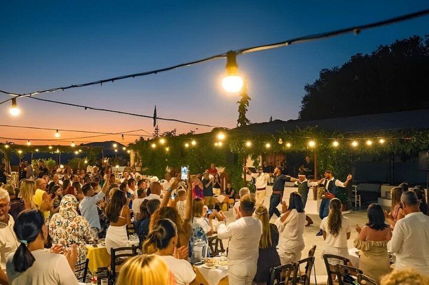 An outdoor gathering with people seated at tables, a stage with performers, and string lights overhead. 2