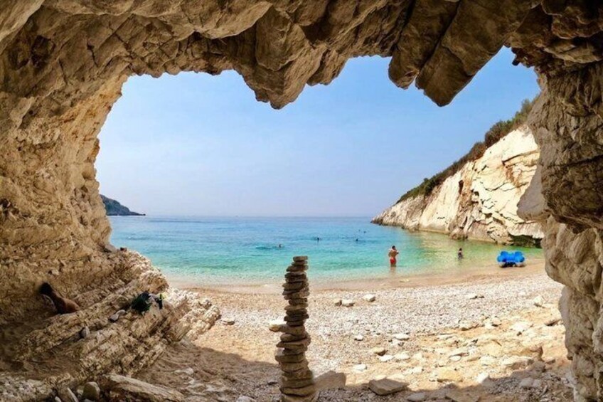 A rocky cave entrance overlooking a beach with clear water and a stack of stones in the foreground. 2