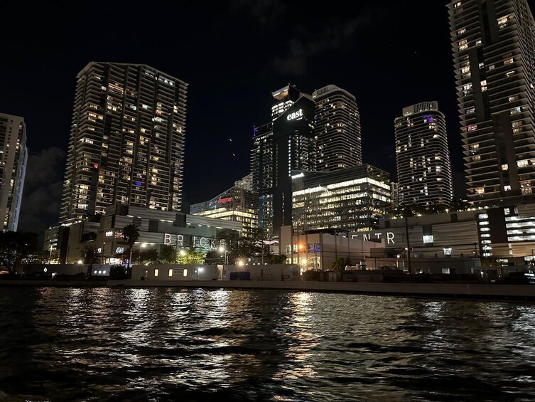 Une vue nocturne d’une ville, avec des gratte-ciel illuminés dont les reflets se mirent dans l’eau. 4