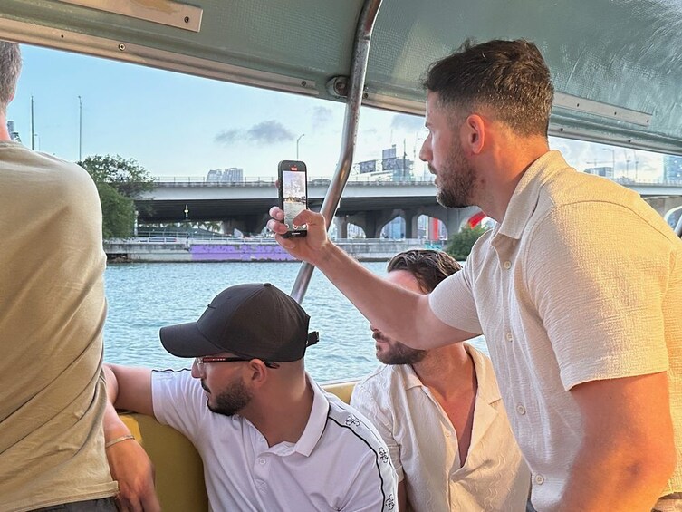 Une personne tenant un smartphone, peut-être en train de prendre une photo ou de filmer une vidéo, avec un pont et de l’eau en arrière-plan. 1