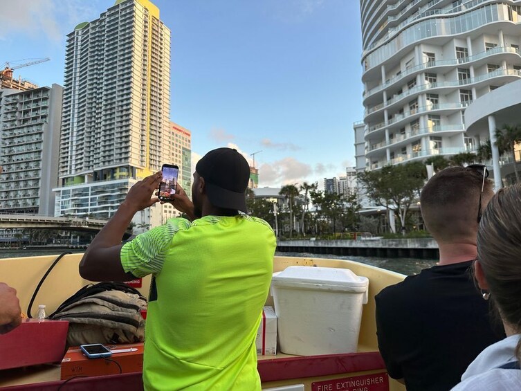 Une personne prenant une photo avec un smartphone, un bateau portant un panneau indiquant la présence d’un extincteur, et des bâtiments modernes en arrière-plan. 3