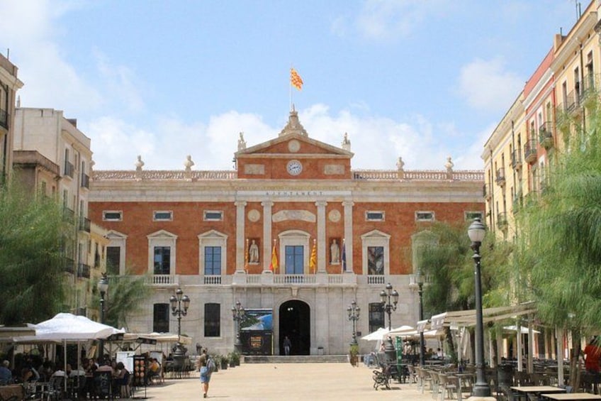 A historic building with a clock, flag, and a plaza with outdoor seating. 3