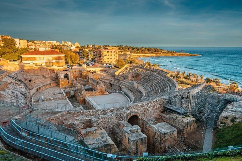 An ancient amphitheater with a partially intact stage and seating area, surrounded by modern buildings and a coastline. 1