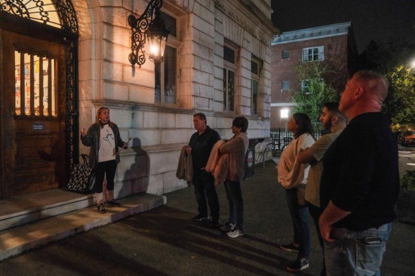 A group of people standing on a sidewalk at night in front of a building with a large door and a street lamp. 4