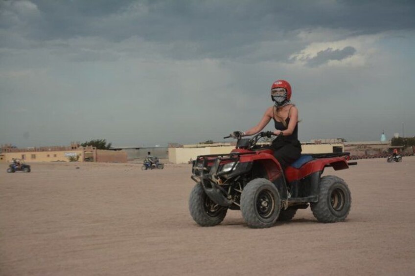 A person riding a red ATV on a sandy terrain with buildings in the background. 3