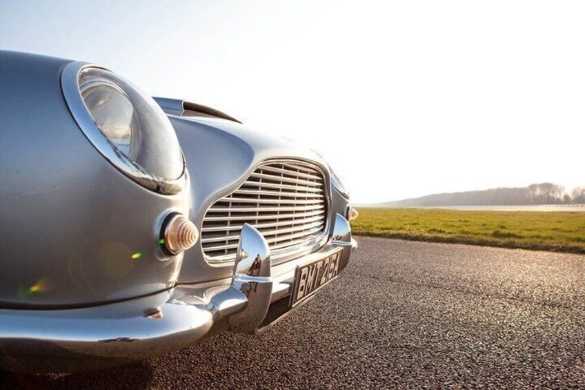 A vintage car with a chrome grille and emblem, parked on a road with a grassy field and trees in the background. 5