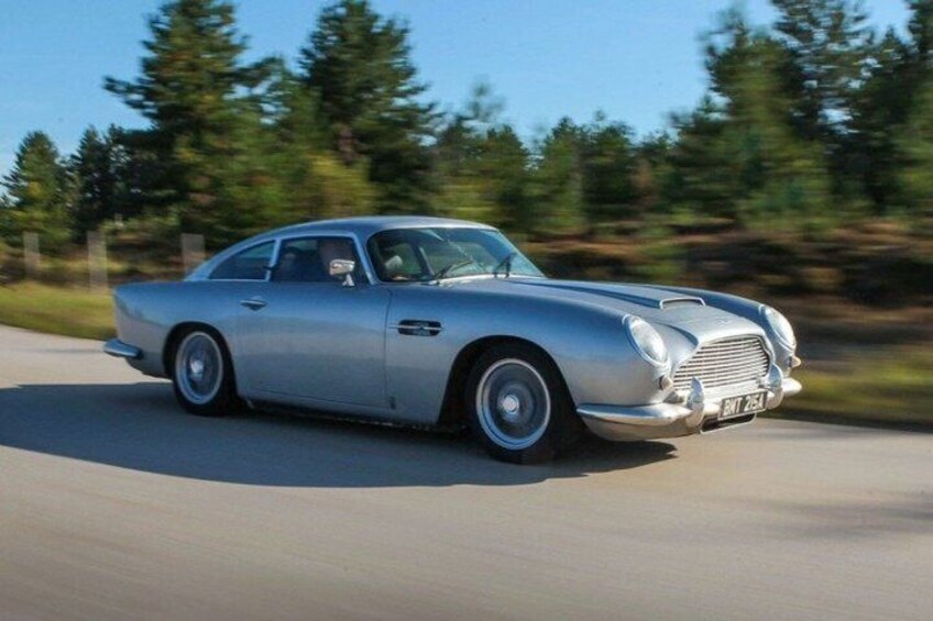A silver vintage car driving on a road with trees in the background. 3