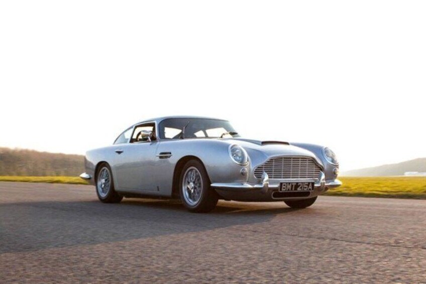 A vintage silver car parked on a paved surface with a grassy field in the background. 2