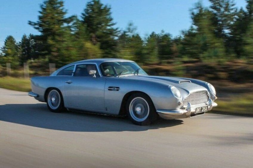A silver vintage car driving on a road with trees in the background. 3