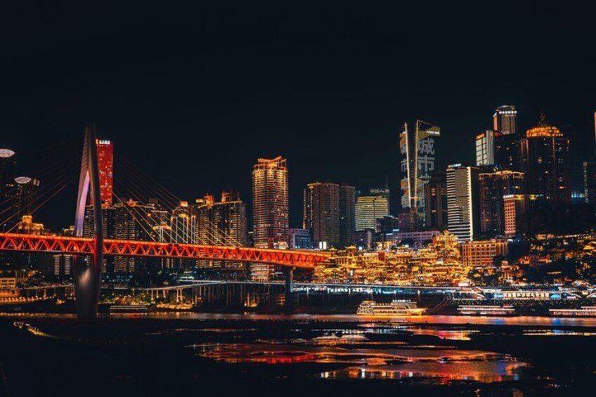 A city skyline at night with illuminated buildings, a prominent red bridge, and a body of water reflecting the lights. 2