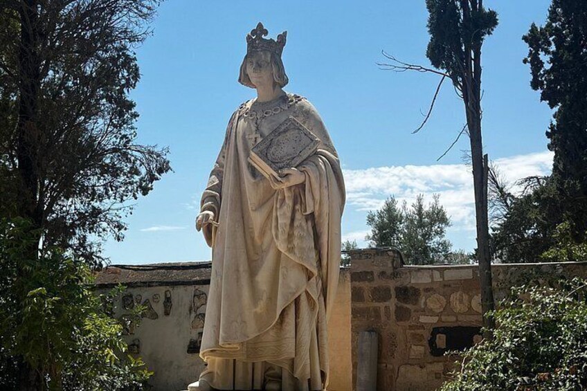 A statue of a historical figure in a robe, standing in a garden with trees and a stone wall in the background. 3