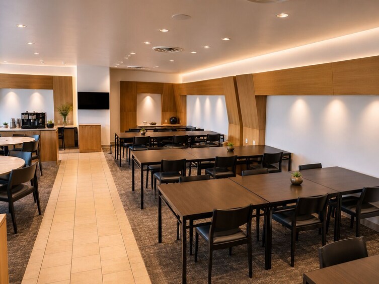A modern dining area with wooden tables and chairs, a kitchen area with a coffee machine, and recessed ceiling lighting. 3
