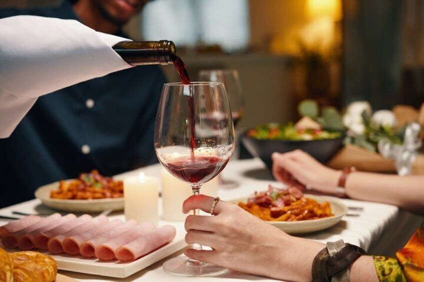 A person pouring red wine into a glass, a plate of sliced ham, and a candle on a table. 1