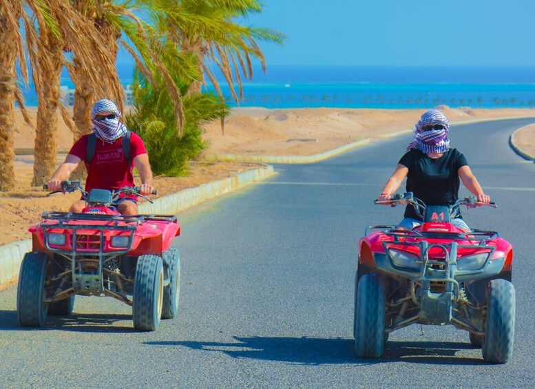 Two people riding ATVs on a road with palm trees and a beach in the background. 2