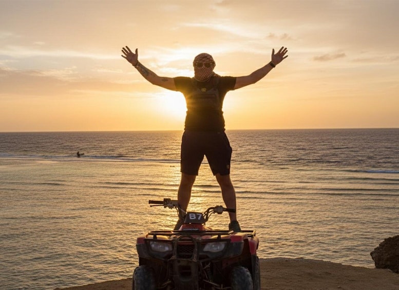 A person standing on an ATV with arms outstretched, facing a sunset over the ocean. 1