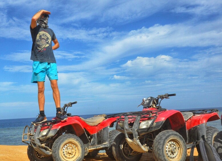 A person standing on a red ATV with a beach and ocean in the background. 4