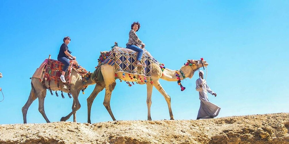 Two camels with riders, one camel is carrying a rider, and a person is walking beside them. 1