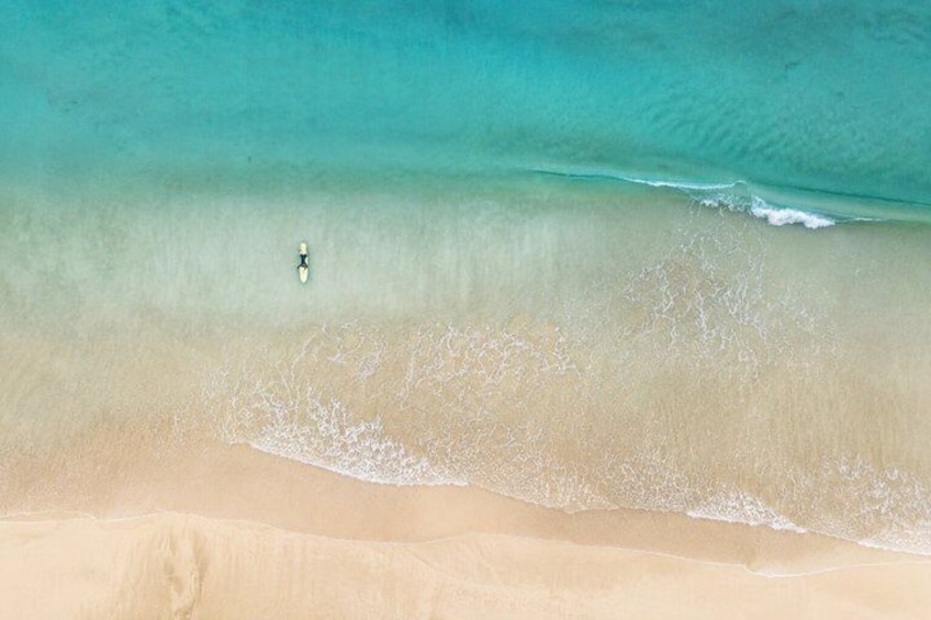 A sandy beach with clear turquoise water and a single person paddling a small boat. 5