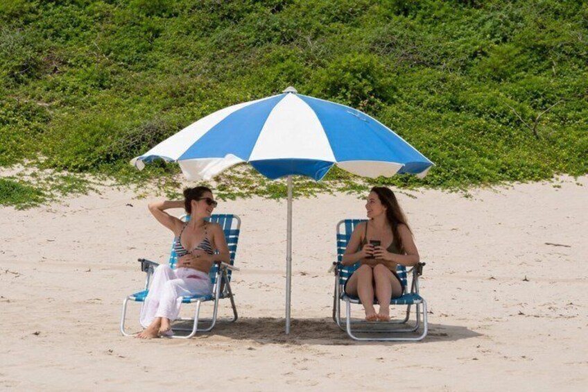 Two people sitting under a blue and white striped beach umbrella on a sandy beach with greenery in the background. 4