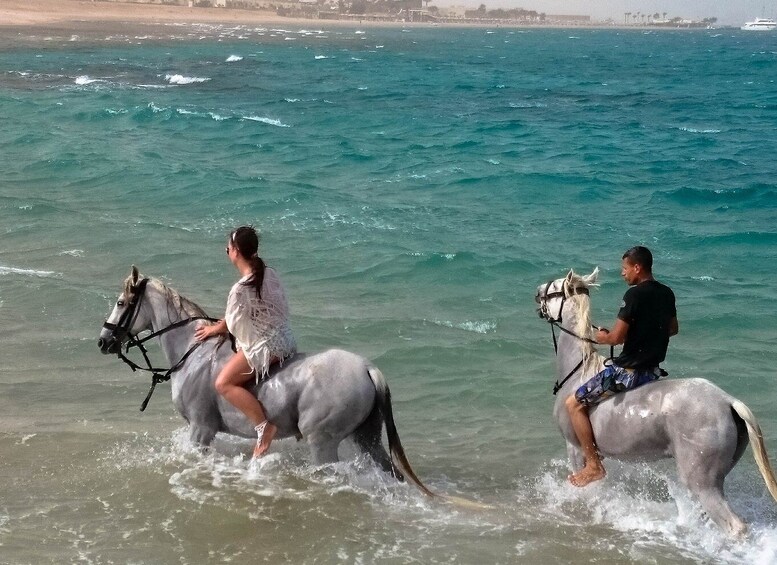 Zwei Personen reiten auf Pferden durch flaches Wasser in der Nähe eines Strandes, im Hintergrund ist eine Stadtansicht zu sehen. 4