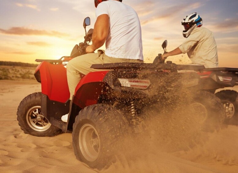 A person riding a red ATV in a sandy area with another person in a white shirt and helmet nearby. 1