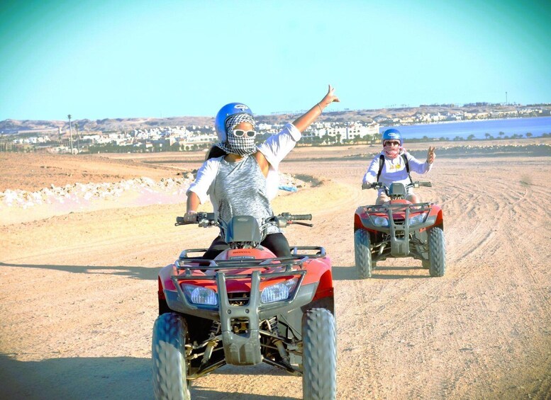 Two people riding ATVs on a dirt road with a beach and buildings in the background. 4