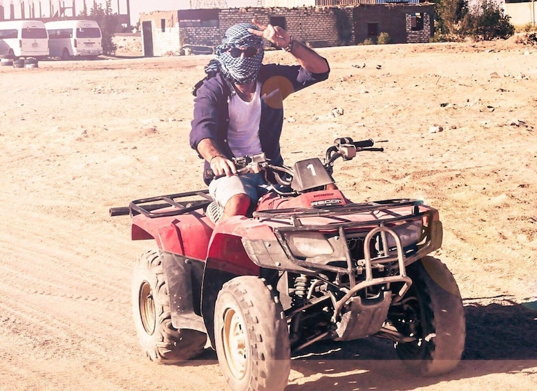 A person on a red ATV in a dusty, open area with buildings in the background. 2
