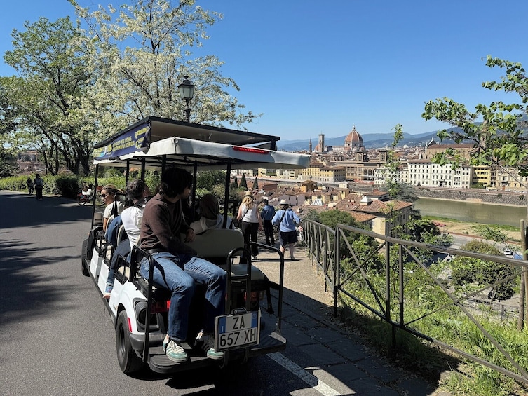 A golf cart with passengers on a paved path, overlooking a river and historic buildings. 1