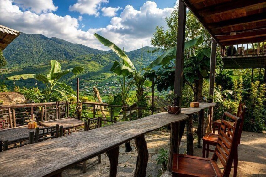 A rustic outdoor dining area with wooden tables and chairs, surrounded by lush greenery and mountains in the background. 2