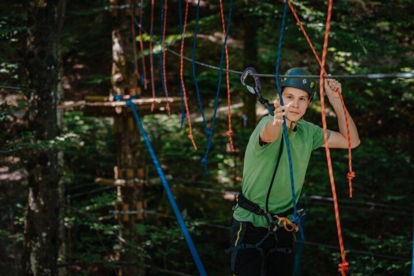 A person engaged in an outdoor activity, wearing a helmet and harness, amidst a forested area with ropes and wooden structures. 2