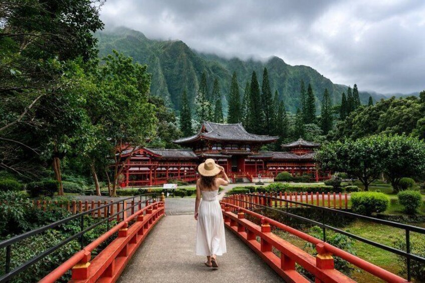 A person in a white dress and straw hat walking on a red bridge towards a traditional building surrounded by greenery and mountains. 4